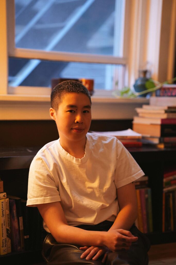 Non Binary Musician in a white tee sitting near their window in their Denver Apartment, posing chill for a photograph