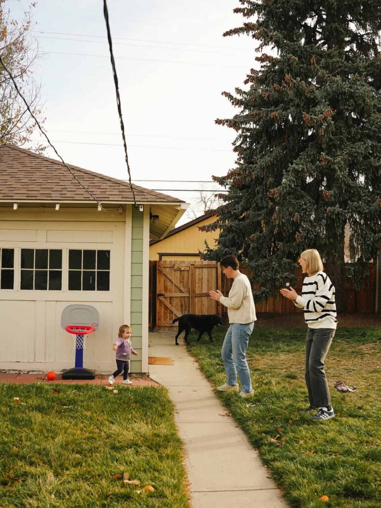 Two proud moms clapping and cheering as their toddler daughter makes a basket in a small backyard hoop while their dog watches during a joyful at-home LGBTQ family session in Denver.
