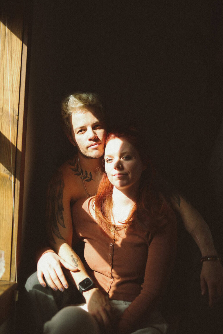 Queer man hugging his fiancé as they sit together by a window during an intimate at-home engagement session near Cherry Creek in Denver.