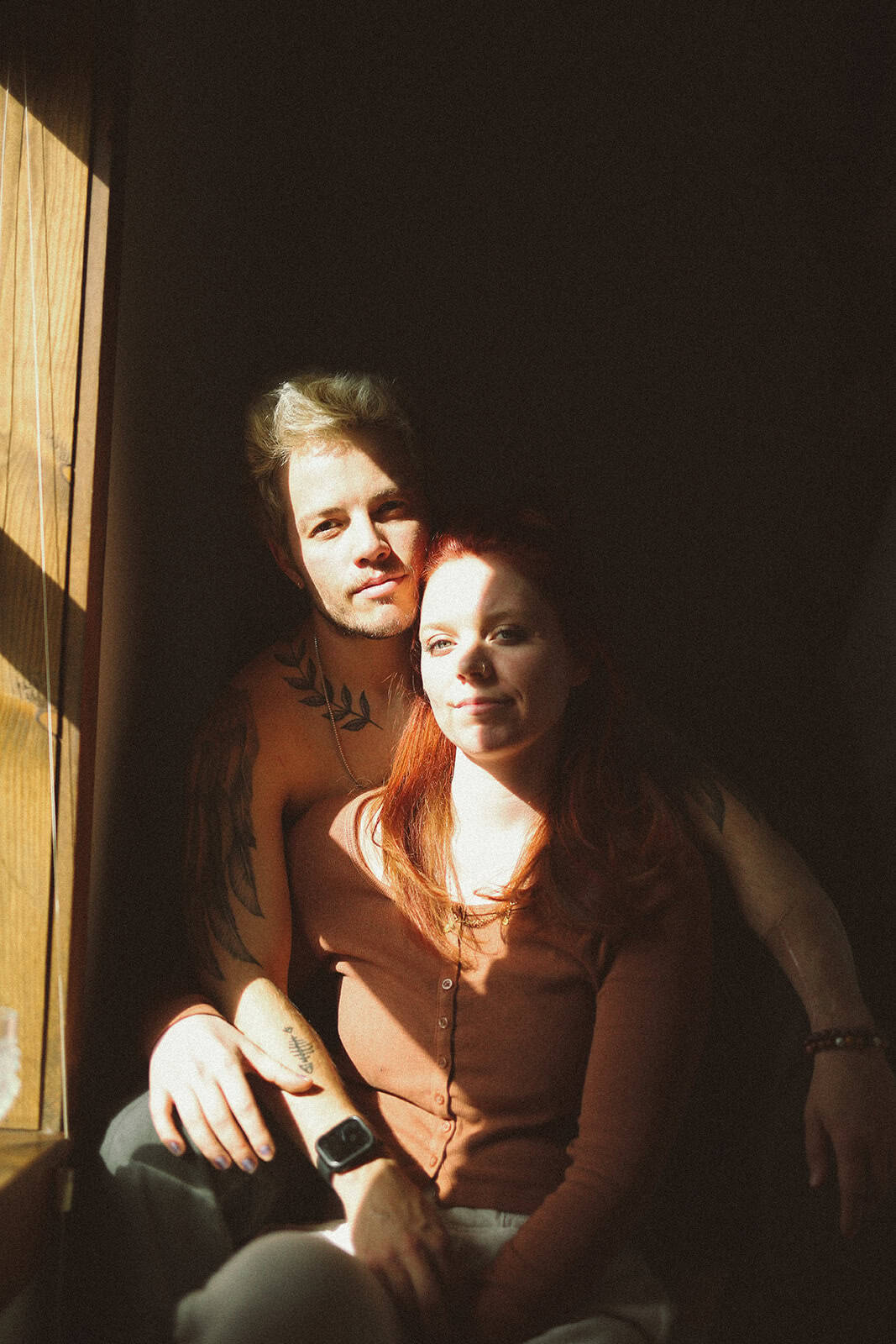 Queer man hugging his fiancé as they sit together by a window during an intimate at-home engagement session near Cherry Creek in Denver.